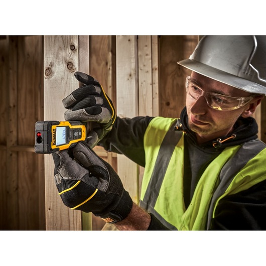 A person wearing a safety vest, work gloves, and a hard hat is using a DEWALT laser distance measurer device to measure the width of wooden planks at a construction site. The person's face is blurred for privacy.