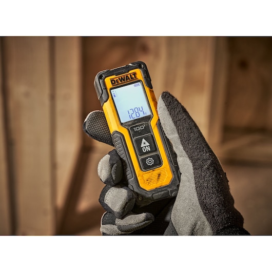 A gloved hand holding a DEWALT laser distance measurer with a digital screen displaying measurement readings, shown in a construction setting with wooden beams in the background.