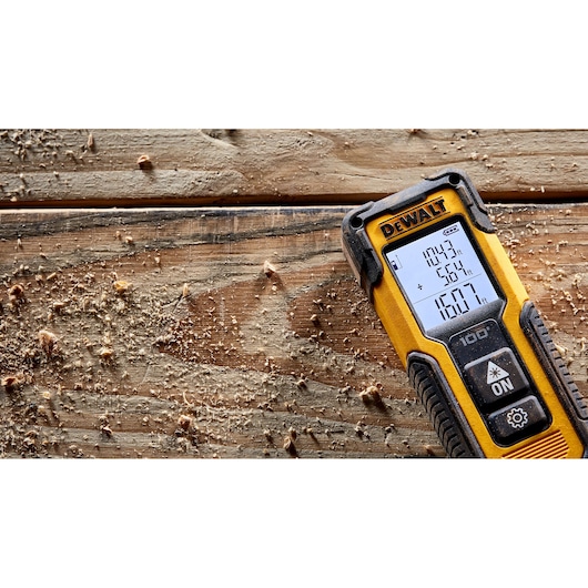 A DEWALT laser distance measurer lies on a wooden surface covered with sawdust. The digital screen displays various measurements. The tool is yellow and black with visible buttons and the DEWALT logo at the top.