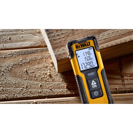 A DEWALT DWHT77100 laser distance measurer resting on a wooden surface with wood shavings, displaying measurements on its screen.