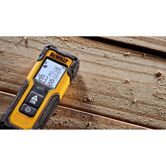 A DEWALT laser distance measurer with a digital screen is placed on a wooden surface covered in sawdust.
