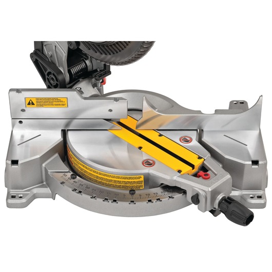 Close-up view of a DEWALT DWS713 miter saw base, showing the metal fence, yellow marked angles, and adjustment knob. The saw is positioned for precise woodworking cuts.