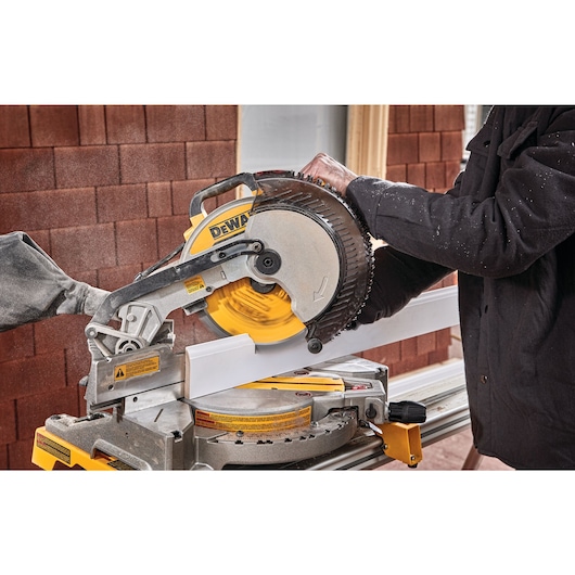 A person using a DEWALT DWS713 miter saw to cut a piece of white trim or molding. The saw is mounted on a workbench and dust is visible in the air.