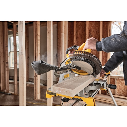 A DEWALT miter saw is being used to cut a wooden board inside a partially constructed room with exposed framing.