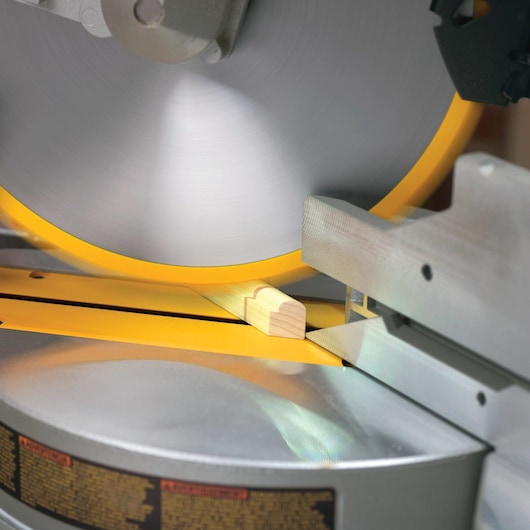 Close-up of a DEWALT miter saw cutting a small piece of wood, showing the spinning blade and metal guide.