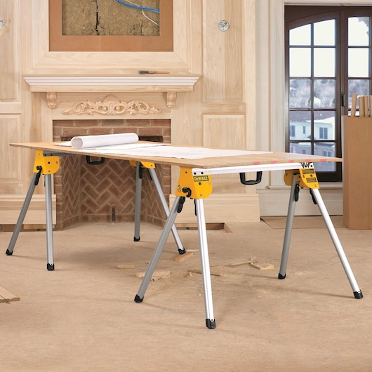 A DEWALT DWS780 yellow and silver sawhorse set used as a work table, with a wooden board and rolled plans on top, placed in a room under renovation with unfinished woodwork and a fireplace.
