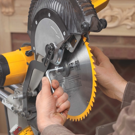 Close-up of a person changing the blade on a DEWALT miter saw, using a wrench to loosen the bolt holding the circular saw blade in place.