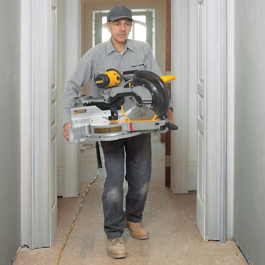 A person carrying a DEWALT DWS780 miter saw in a hallway during construction or renovation. The individual is wearing work clothes and boots, with the face blurred for privacy.