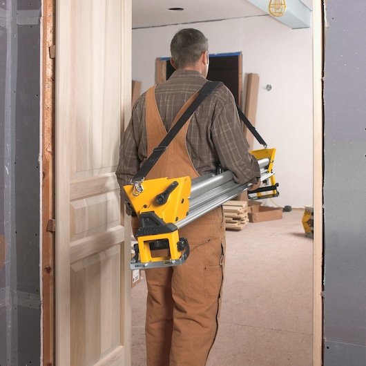 A person wearing brown overalls and a plaid shirt is carrying a DEWALT miter saw stand through a doorway in a room under construction.