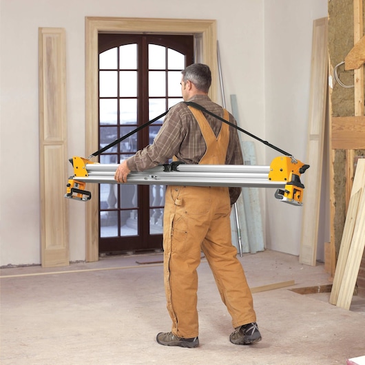 A person wearing brown overalls carries a DEWALT DWS780 photography equipment support rail with yellow ends and a strap in a room under construction.