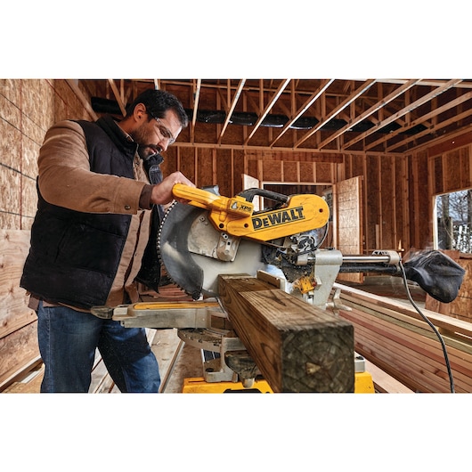 A person operating a DEWALT DWS780 miter saw in a construction site, cutting a wooden beam inside a partially built structure.