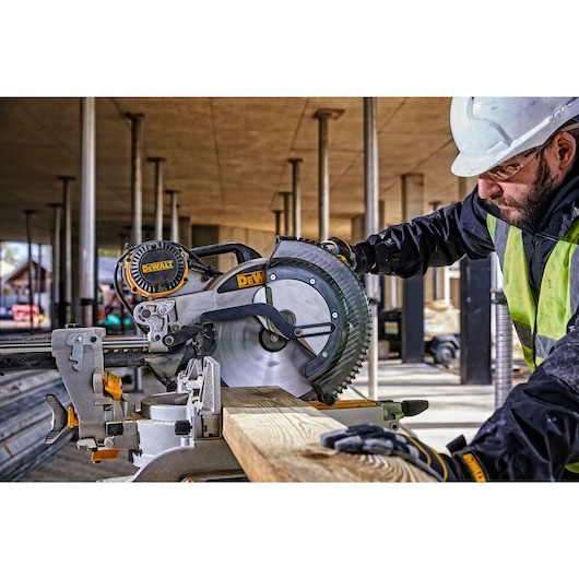 A DEWALT DWS780 miter saw is being operated by a worker wearing safety gear and a helmet at a construction site. The saw is cutting a piece of wood.