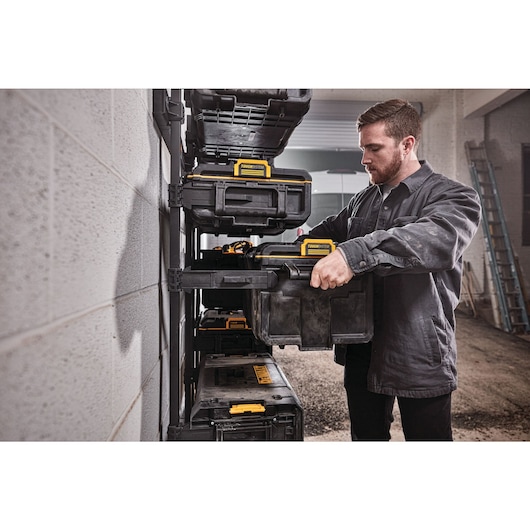 A person is organizing DEWALT black and yellow tool storage boxes on a wall-mounted shelving unit in a workshop.