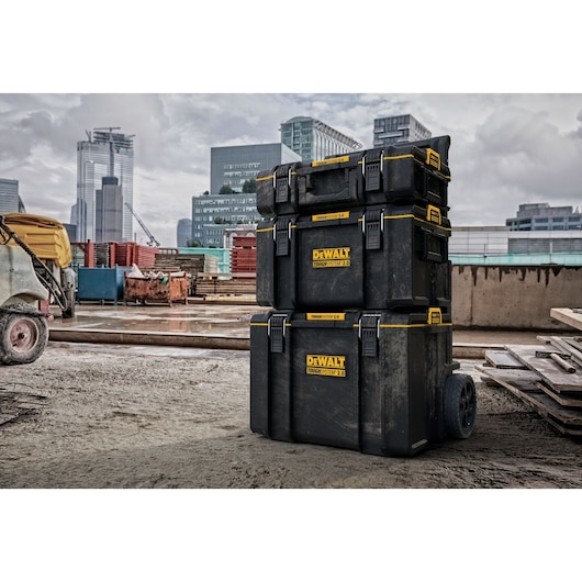 Stack of three DEWALT ToughSystem 2.0 tool storage boxes with wheels, shown on a construction site with modern buildings in the background.
