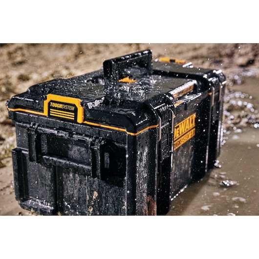 A DEWALT ToughSystem black and yellow toolbox sitting on wet muddy ground, being splashed with water, showcasing its durability and waterproof design.