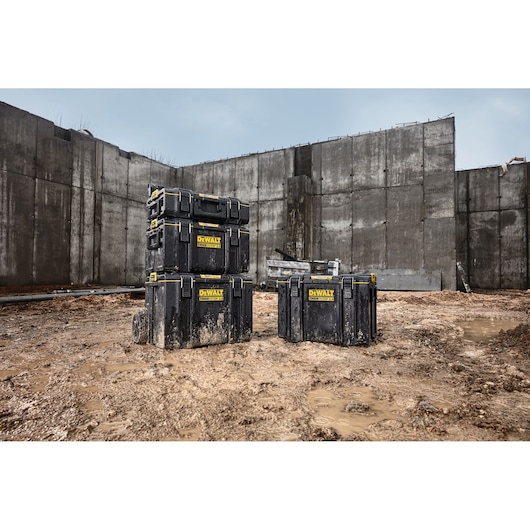 Three DEWALT ToughSystem tool storage boxes are shown outdoors on a muddy construction site, positioned in front of large concrete walls. The boxes are black with yellow DEWALT logos and vary in size.