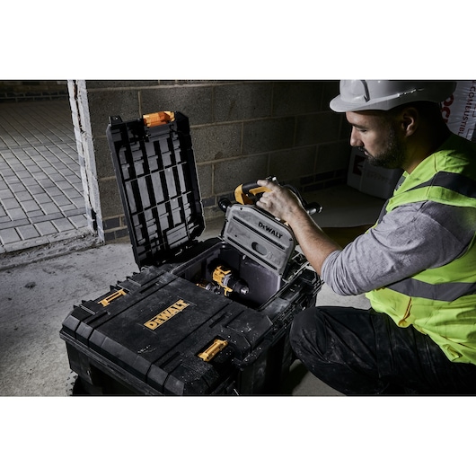 A person wearing a high-visibility vest and a safety helmet is handling a tool inside an open DEWALT storage box, placed on a concrete floor at a construction site. The person's face is intentionally blurred for privacy.