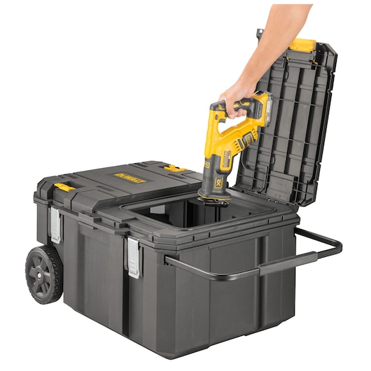 A person is placing a yellow and black DEWALT power tool into a large black wheeled toolbox with an open lid and a pull handle.