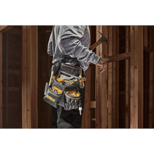 Person wearing a DEWALT tool belt loaded with various hand tools and construction accessories, standing at a wooden construction site and holding a hammer.