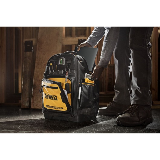 A DEWALT tool backpack with multiple compartments and organized tools, being used at a construction site. A person is placing a tablet into the backpack.