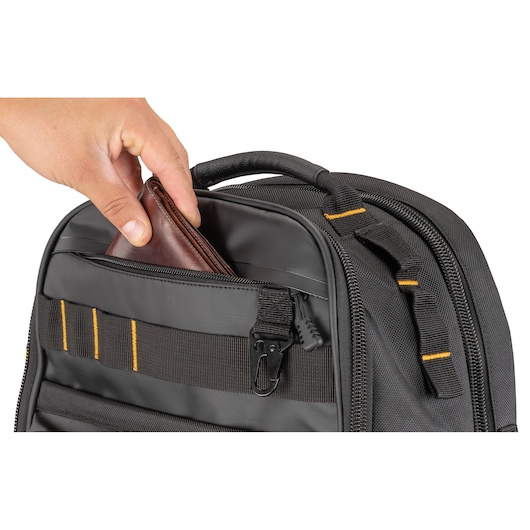 A hand is placing a brown leather wallet into the front zippered pocket of a black DEWALT backpack with yellow stitching.