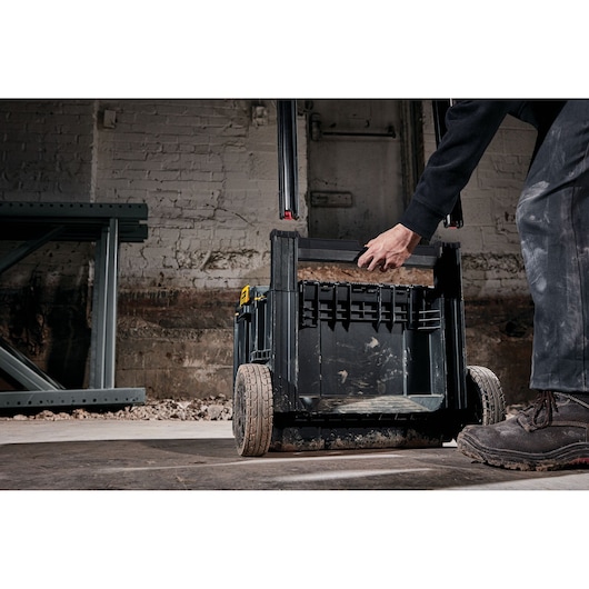 A person is using a DEWALT rolling toolbox with large wheels in an industrial setting. The toolbox is black with a sturdy handle and appears to be used for construction or worksite storage.