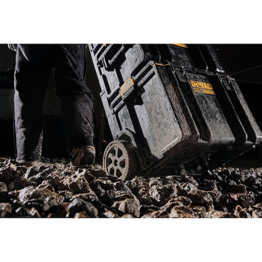 A person pulls a DEWALT ToughSystem tool storage box with large wheels over rough, rocky ground. The tool box and the person's pants are covered in dirt, highlighting durability for tough job site conditions.