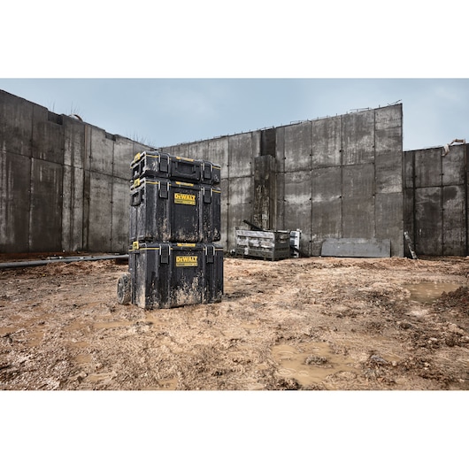 Three stacked DEWALT toolboxes covered with dirt and mud are shown on a muddy construction site with concrete walls in the background.